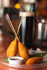 Corn dogs on sticks with chicken sausage, deep fried in egg batter, served with balsamic vinegar and a glass of cold stout, on bokeh background