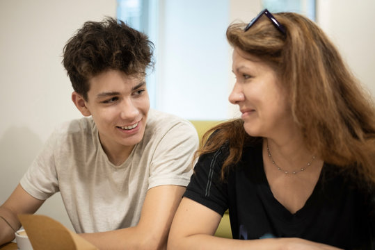 The Teenager Communicates With His Mom In The Cafe And Smiles. Mom Looks At Her Son With Love And Does Not Believe That He Grew So Fast. Focus On The Guy