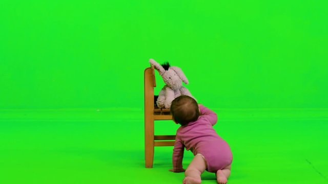 A baby girl crawls, stands using a chair, looks towards the camera and laughs, over a green screen.