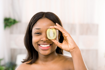 Funny smiling african american young woman holding kiwi half in front of her eye over white...