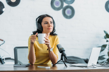 pensive radio host looking away while sitting at workplace and holding coffee cup