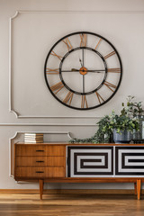 Real photo of a large, metal clock hanging on a white wall with molding above a wooden cupboard in...