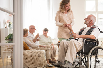 Senior man on wheelchair with helpful nurse holding his hand and friends sitting on couch drinking...