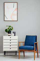 Poster above white cabinet with plant next to blue wooden armchair in grey flat interior. Real photo