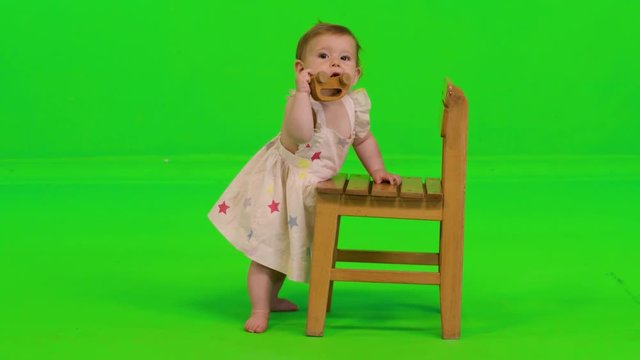 A baby girl plays with a toy, smiles and laughs over a green screen, wearing a dress.