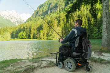young man in a wheelchair fishing at the beautiful lake on a sunny day, with mountains in the back