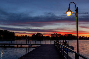 Piękny zachód słońca nad jeziorem Kalwa na Mazurach w Polsce., na pierwszym planie pomost na jeziorze i lampa.