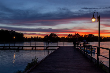 Piękny zachód słońca nad jeziorem Kalwa na Mazurach w Polsce., na pierwszym planie pomost na jeziorze i lampa.