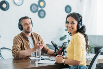 cheerful radio host looking at camera while sitting at workplace near colleague