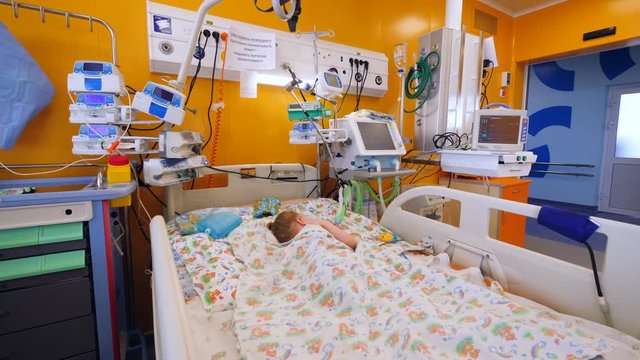 Sick child sleeps in bed in a hospital ward with medical machines.