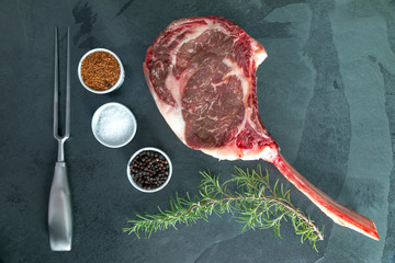 Raw, dry-aged wagyu tomahawk steak, salt, pepper, bbq rub and rosemary on a  black stone table. Top...