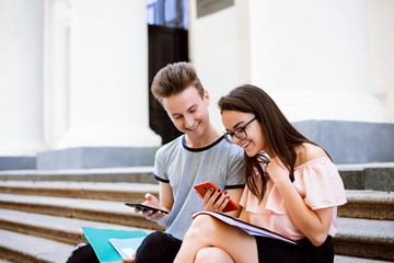 Happy students sitting on stairs of college using mobile phones, texting, taking pictures and...