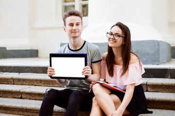 Smiling man and woman using modern laptop convertible computer showing screen to the camera sitting on stairs of old university. Using technology in studying