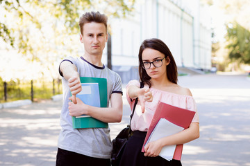 Two angry and bewildered friends holding books, notes and other learning materials gesturing thumbs down in the street near old conventional university