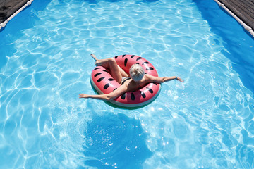 Woman with short hair swimming relaxing in a pool with pink floatie Inflatable doughnut, blue water, chill, tanning under sun.