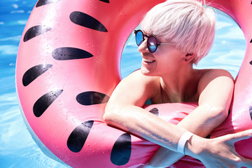 Portrait of woman with short blonde hair swimming relaxing in a pool with pink floatie Inflatable doughnut, blue water, chill, tanning under sun.