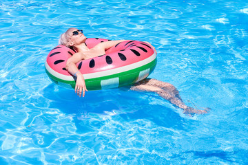 Woman with short blonde hair swimming relaxing in a pool with pink floatie Inflatable doughnut, blue water, chill, tanning under sun.