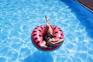 Woman with short hair swimming relaxing in a pool with pink floatie Inflatable doughnut, blue water, chill, tanning. Space for text layout.