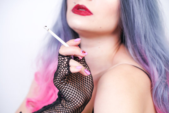 bad girl with piercing and long grey pink hair in black corset and torn fishnet pantyhose with mesh gloves sitting on the floor and Smoking cigarette on white background in Studio alone