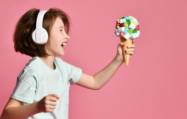 Happy teen girl kid walks in headphones sings with a big ice cream in waffles cone with tasty toppings give it on pink