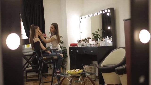 Beautiful female makeup artist doing makeup for a young redhead girl in a beauty salon sitting in front of a large mirror. Concept of preparation for the holiday and meeting