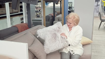 Happy senior woman choosing cushions for her living room at furniture store. Elderly female customer sitting on a couch on sale, smiling to the camera. Lifestyle, retirement concept