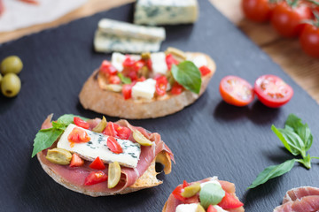 Traditional Italian bruschetta with blue cheese, feta, tomatoes, basil leaves, jamon on black stone...