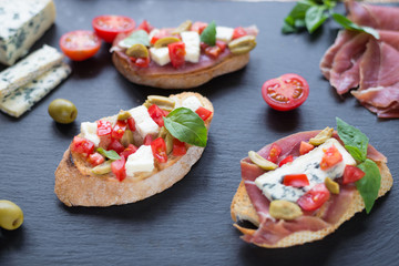 Traditional Italian bruschetta with blue cheese, feta, tomatoes, basil leaves, jamon on black stone...