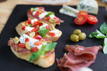 Traditional Italian bruschetta with blue cheese, feta, tomatoes, basil leaves, jamon on black stone background.