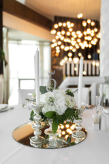 Wedding table setting is decorated with fresh flowers and white candles. Wedding floristry. Bouquet with roses, hydrangea and eustoma. On the background blur are burning garlands with light bulbs.