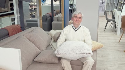 Cheerful elderly man trying soft cushions on sale at furniture store. Happy senior man sitting on a couch at furnishings store. Elderly male customer at the furniture store