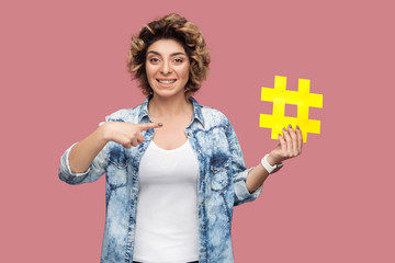Portrait of happy young woman in blue shirt with curlty hair holding large big yellow hashtag sign and pointing finger, social media concept. Indoor, isolated, studio shot, copy space, pink background