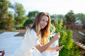 Elegant charming woman texting romantic message on mobile phone. Portrait of luxary lady in glasses enjoying summer sunset in cafe outdoor. Communicating by smartphone in travel..