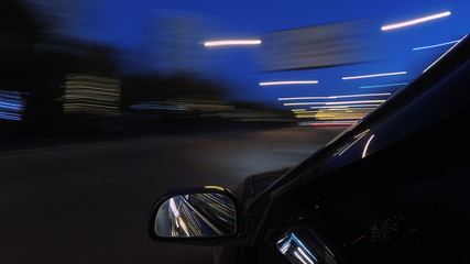 The dark-colored car is moving fast on the illuminated road of the night city, on a blurry flying background one can see a part of the car with a mirror