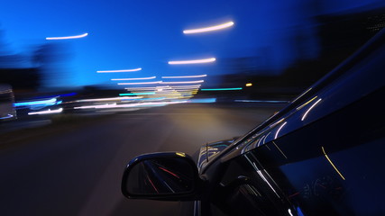 The dark-colored car is moving fast on the illuminated road of the night city, on a blurry flying background one can see a part of the car with a mirror