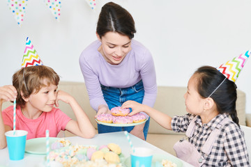 Little girl in birthday cap taking donut from plate held by mother