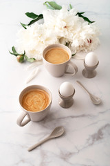 Boiled eggs in ceramic egg cups, two cups of coffee with ceramic spoons on background of beautiful white peony. Breakfast concept. Copy space.