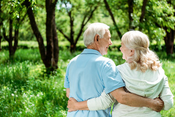 smiling pensioner hugging happy senior wife while standing in green park