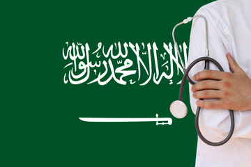 male doctor in white coat holding in his hand a medical instrument with a stethoscope against the background of the national flag Saudi Arabia, close-up, concept, copy space