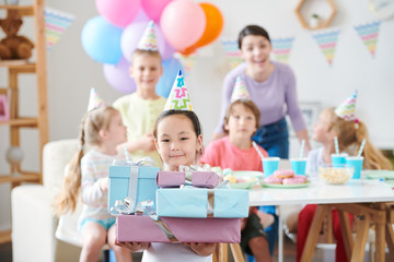 Little Asian girl with stack of birthday gifts looking at you at party