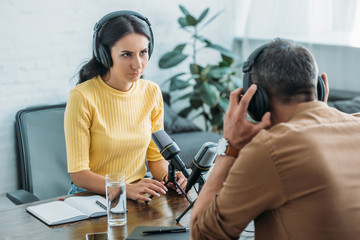 attractive serious radio host looking at colleague while sitting at workplace in studio