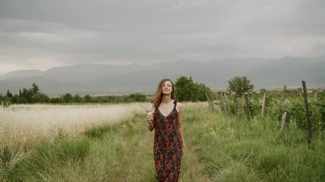Young beautiful woman drinking wine from a glass and walking along in the fields and vineyards with mountains in the background. 4k.