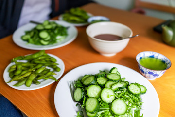 Home with wooden table and green salad dish with Japanese cucumbers and mizuna greens and boiled edamame soy beans with tea