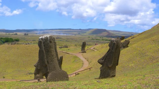 AERIAL: Unrecognizable tourists walk along the scenic path leading past a volcano and big moai sculptures on Easter Island. Spectacular view of legendary statues scattered around the green meadows.