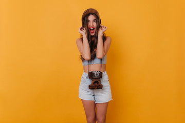 Pretty female photographer in white denim shorts fooling around in studio. Indoor portrait of graceful brown-haired girl in stylish hat posing with camera.