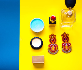 Modny kolaż zawierający ręcznie szyte kolczyki sutasz, szminkę, perfumy oraz kolorowe kosmetyki w ujęciu top view na żółto niebieskim tle - obrazy, fototapety, plakaty
