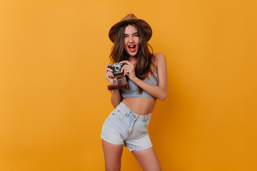 Excited female photographer standing in confident pose on yellow background. Studio shot of beautiful brunette girl in stylish hat and white shorts holding camera and laughing.