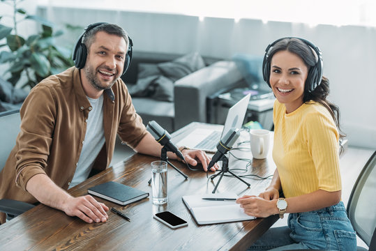 two cheerful radio hosts smiling at camera while sitting at workplace in broadcasting studio