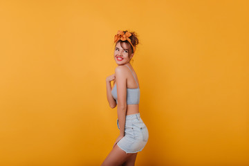 Happy caucasian girl in summer outfit looking away while posing in studio. Portrait of enchanting young woman in retro attire isolated on yellow background.