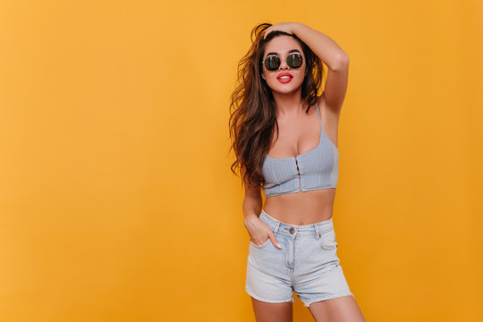 Happy girl in vintage denim shorts standing in confident pose on yellow background. Indoor portrait of enchanting young lady in summer casual outfit plays with her hair.